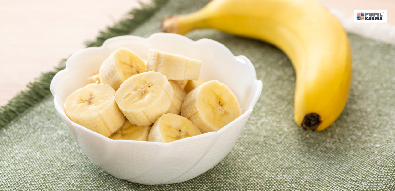 Czy pies może banany? Najczęstsze błędy w podawaniu bananów psom. Biała miska z pokrojonym w kosteczkę bananem, obok cały banan w skórce. W prawym górnym rogu logotyp sklepu pupilkarma - polskiego producenta karmy dla psów i kotów.